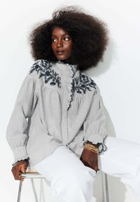 Chemise à rayures blanche et marine, ornée de broderies florales sur les épaules, avec un col volanté et des poignets froncés ; assortie à un pantalon blanc.
