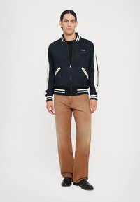 Veste bomber zippée marine avec des rayures blanches sur les manches, associée à un pantalon large marron clair et des chaussures noires. Fond uni.