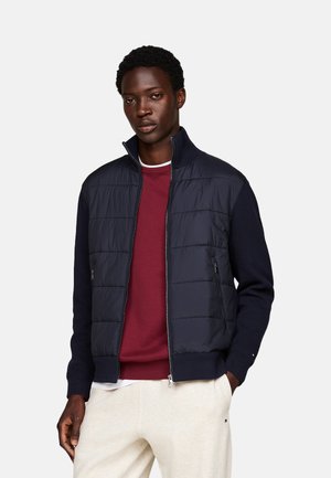 Chaqueta de punto azul marino con cuello alto, bolsillos con cremallera y un material de punto en las mangas que contrasta, combinada con una camiseta de cuello redondo burdeos.