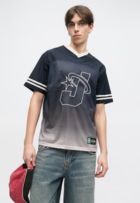 Ναυτικό μπλε αθλητικό μπλουζάκι από διχτυωτό ύφασμα με εφέ βαθμιδωτής μετάβασης σε γκρι, διαθέτει λευκό λογότυπο "J" και ρίγες στα μανίκια. Φοριέται με ανοιχτού χρώματος τζιν.