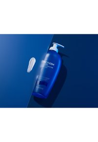 Blå plastflaske med en hvit pumpe merket "BIOTHERM ULT-CORRECTIVE BODY MILK." En klatt med hvit krem ligger ved siden av den på en blå bakgrunn.