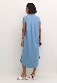 Vestido de mezclilla sin mangas, azul claro, hasta la rodilla con aberturas laterales y un dobladillo redondeado. Presenta un cuello y sutiles detalles de costura.