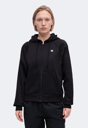Ung person med kort blondt hår, iført en sort zip-up hættetrøje med et lille Champion-logo på brystet, mod en ensfarvet baggrund.