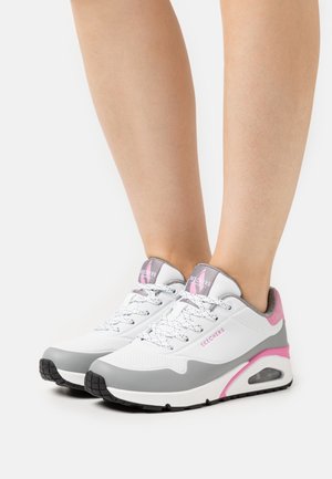 Jambes portant des baskets Skechers blanches, grises et roses avec des lacets blancs, debout sur une surface blanche sur un fond uni.