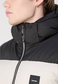 Sort og off-white polstret jakke med høj krave, lynlåslukning og Calvin Klein-branding på stofmærket. Blød tekstur, moderne design.