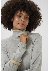 Světle šedý rolák vyrobený z pleteného materiálu, s vroubkovaným límcem a manžetami. Na levém zápěstí je nošena béžová náramková bangle.