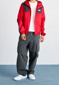 Czerwona i czarna kurtka z kapturem, wykonana z lekkiego materiału, z logo na piersi i regulowanymi mankietami; zestawiona z szarymi spodniami cargo.