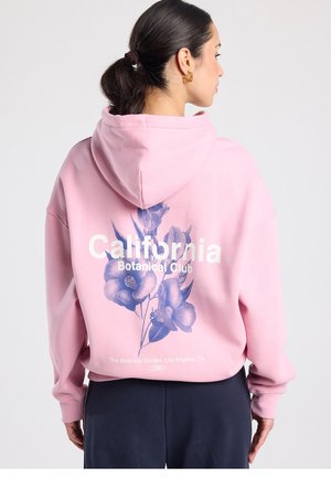 Sweat à capuche rose avec un grand motif floral bleu, texte "California Botanical Club" et capuche avec cordon de serrage. Matière douce, coupe décontractée.