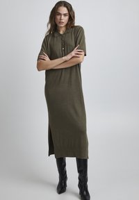 Robe polo en maille verte olive, à manches courtes, avec une coupe droite, une fente sur le côté et un détail de bouton au col, assortie à des bottes noires au genou.