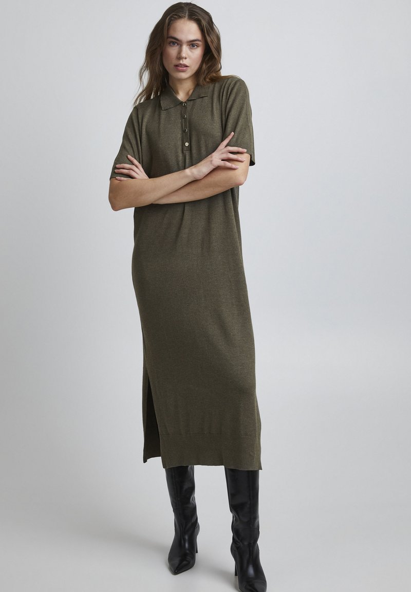 Rochie polo tricotată, cu mânecă scurtă, de culoare verde măslin, cu croială dreaptă, slit lateral și detalii cu nasturi la guler, asortată cu cizme negre până la genunchi.