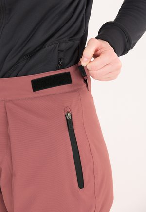 Pinkfarbene Hosen mit strukturierter Oberfläche, ausgestattet mit einer schwarzen Reißverschlusstasche und einem mit Klettverschluss verstellbaren Bund. Die Hand befindet sich am Bund.