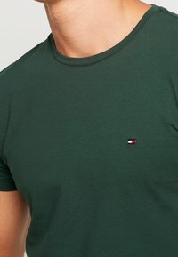 Mand iført en mørkegrøn T-shirt med rund hals og et lille, rektangulært logo i rød, hvid og marineblå på venstre bryst.