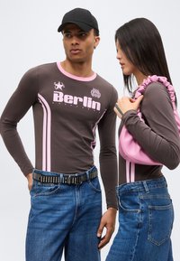 Άνδρας και γυναίκα φορούν ταιριαστά γκρι και ροζ μακρυμάνικα μπλουζάκια και μπλε τζιν; η γυναίκα κρατάει μια ροζ τσάντα με πιέτες στον ώμο.