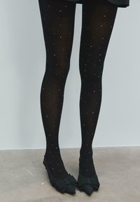 Czarne rajstopy w wzory z rozrzuconymi srebrnymi kropkami, połączone z czarnymi płaskimi butami z dużymi kokardami i spiczastymi czubkami.