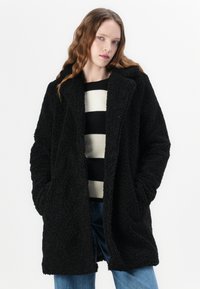 Black fluffy coat with a soft texture, featuring a notched collar and side pockets, layered over a striped black and white sweater.