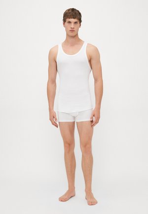 Junger Mann, der barfuß steht und ein weißes geripptes Tanktop sowie passende weiße Boxer-Shorts trägt, vor einem schlicht weißen Hintergrund.