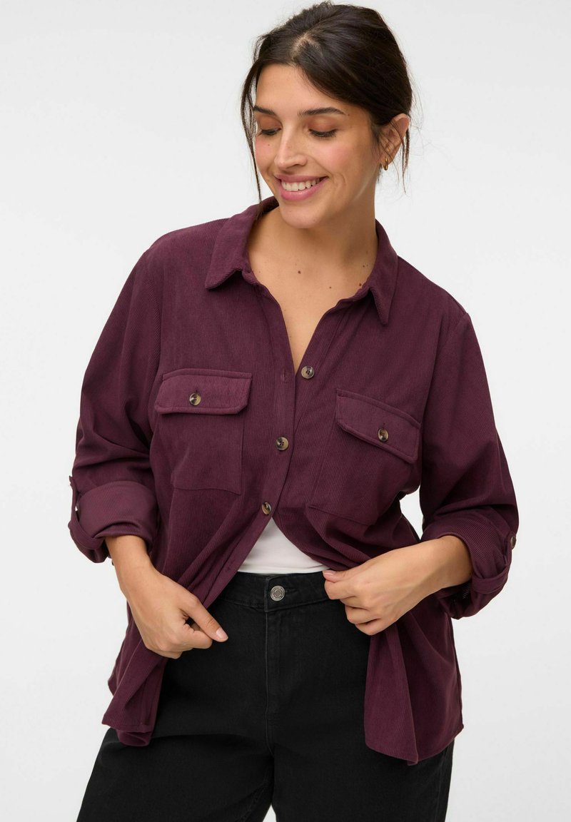 Femme souriante ajustant une chemise en velours côtelé violette déboutonnée par-dessus un top blanc et un pantalon noir sur un fond uni.