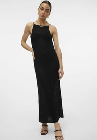 Abito lungo nero in maglia con spalline sottili, motivo testurizzato, fessure laterali e una silhouette aderente. Indossato con sandali neri con cinghie.