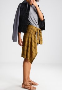 Svart och grått färgblockerat jacka över en grå t-shirt, parat med en senapsgul kjol med rutmönster och bruna sandaler med remmar.