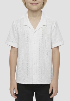 Niño con una camisa blanca de manga corta abotonada con rayas verticales de ojales y pantalones negros, de pie sobre un fondo neutro.