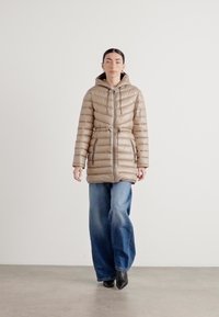 Cappotto imbottito beige con cappuccio, vita con coulisse, cuciture orizzontali e tasche con zip, abbinato a jeans a gamba larga e stivali alla caviglia.