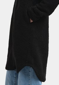 Svart, texturerad fleecejacka med lös passform, sidfickor och rundad nederkant. Kombinerad med ljusblå denimjeans.