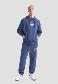 Felpa in cotone blu navy con pantaloni abbinati, dotata di una tasca frontale, caratterizzata da un logo grafico testuale giallo e polsini a costine alle caviglie.
