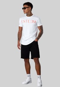 Weißes T-Shirt mit dem Aufdruck "ESTEEM" in leuchtendem Orange, kombiniert mit schwarzen Denim-Shorts. Getragen mit weißen Turnschuhen und schwarzen Sonnenbrillen.