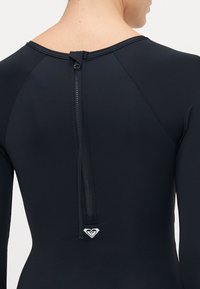 Close-up of a person wearing a black long-sleeve wetsuit with a vertical back zipper and a small white logo near the zipper end.