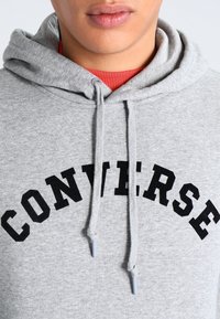 Gray hoodie featuring a black "CONVERSE" logo in an arched design. Material appears soft, with adjustable drawstrings and a classic kangaroo pocket.