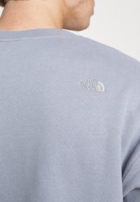 Světle modrá mikina vyrobená z měkké tkaniny, s kulatým výstřihem a žebrovanými manžetami. Vyšité logo na horní části zad, minimalistický design.