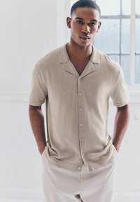 Beige Hemd mit kurzen Ärmeln und Knopfverschluss, hergestellt aus leichtem Stoff mit einer subtilen Textur, mit einem Schalkragen und fünf Knöpfen auf der Vorderseite.