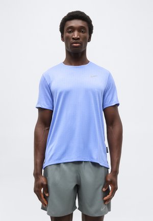 Mand står iført lyseblå kortærmet sportstrøje og grå shorts mod en ensfarvet hvid baggrund.