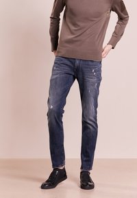 Jeans de denim azul oscuro con un ajuste ligeramente ajustado, que presentan detalles desgastados y una textura suave, combinados con zapatillas bajas negras.