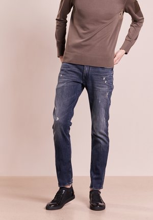 Mörkblå denimjeans med en något smal passform, med slitna detaljer och en mjuk textur, kombinerade med svarta låga sneakers.
