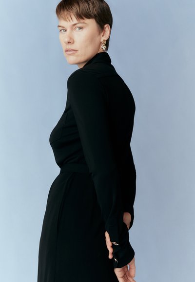 Black long-sleeve dress with a fitted silhouette and belt detail, complemented by a round gold earring. Solid color against a soft blue background.