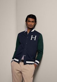 Marineblauwe en groene varsityjas met witte accenten, voorzien van een grote "H" op de borst en een gestreepte geribbelde zoom. Gecombineerd met een geruite shirt.