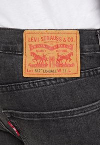Svarta denimjeans med en gul-orange läderlapp. Lappen har röd text och grafik av hästar och byxor. Modell Lot 512, W 31, L 32.