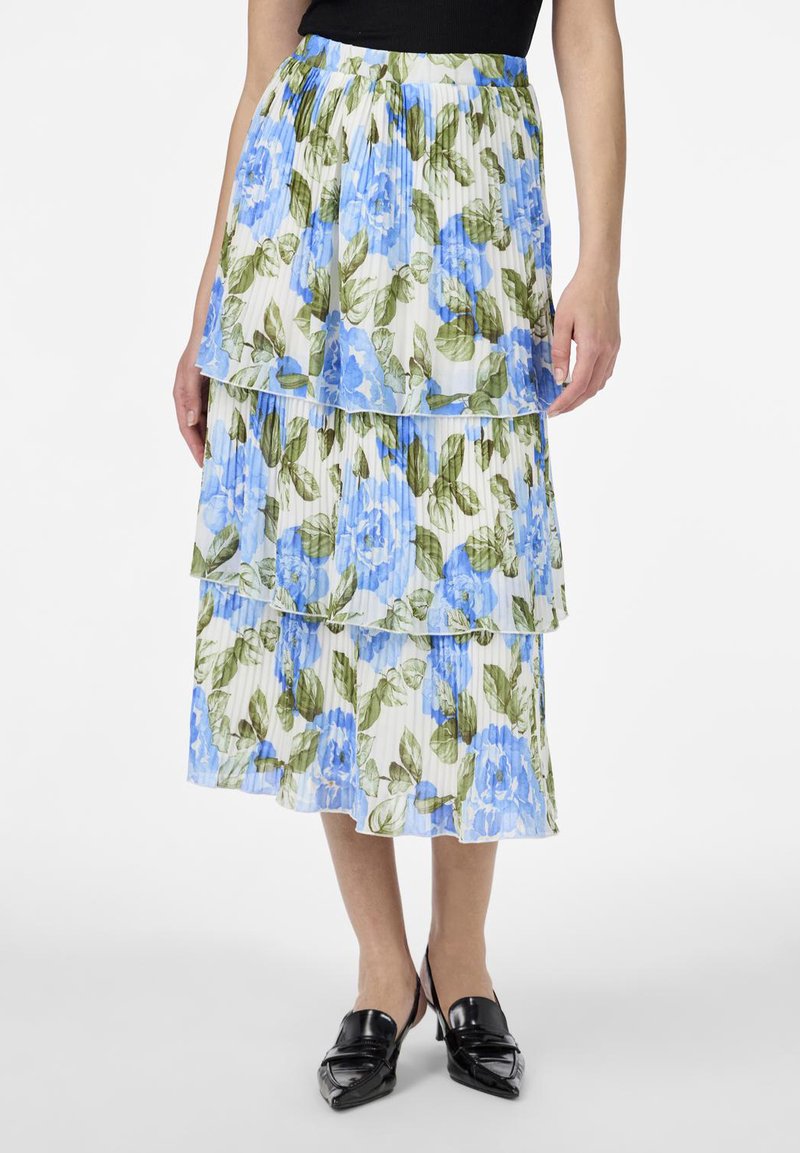 Jupe plissée avec trois niveaux superposés ornés de motifs floraux bleus sur fond blanc et de feuilles vertes. Le matériau semble léger.