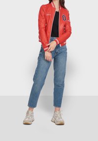 Kobieta ubrana w czerwoną kurtkę bombowiec, czarną bluzkę, niebieskie dżinsy i białe trampki, stojąca na tle czystej białej ściany.