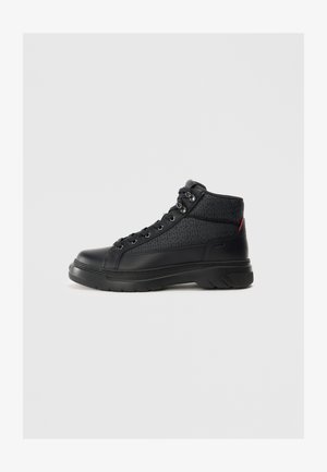 Sneaker alta nera con tomaia in pelle liscia, accenti in tessuto a motivo, occhielli in argento e una spessa suola in gomma. Dettaglio rosso sul tallone.