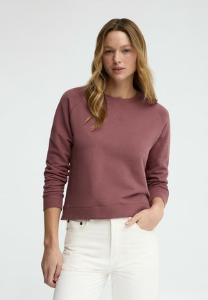 Donna con capelli lunghi e ondulati, che indossa una felpa malva a maniche lunghe e pantaloni bianchi, in piedi con una mano in tasca davanti a uno sfondo neutro.