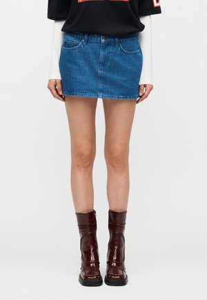 Donna con minigonna di jeans blu, camicia nera a maniche lunghe con maniche bianche e stivali lucidi marrone scuro fino a metà polpaccio.