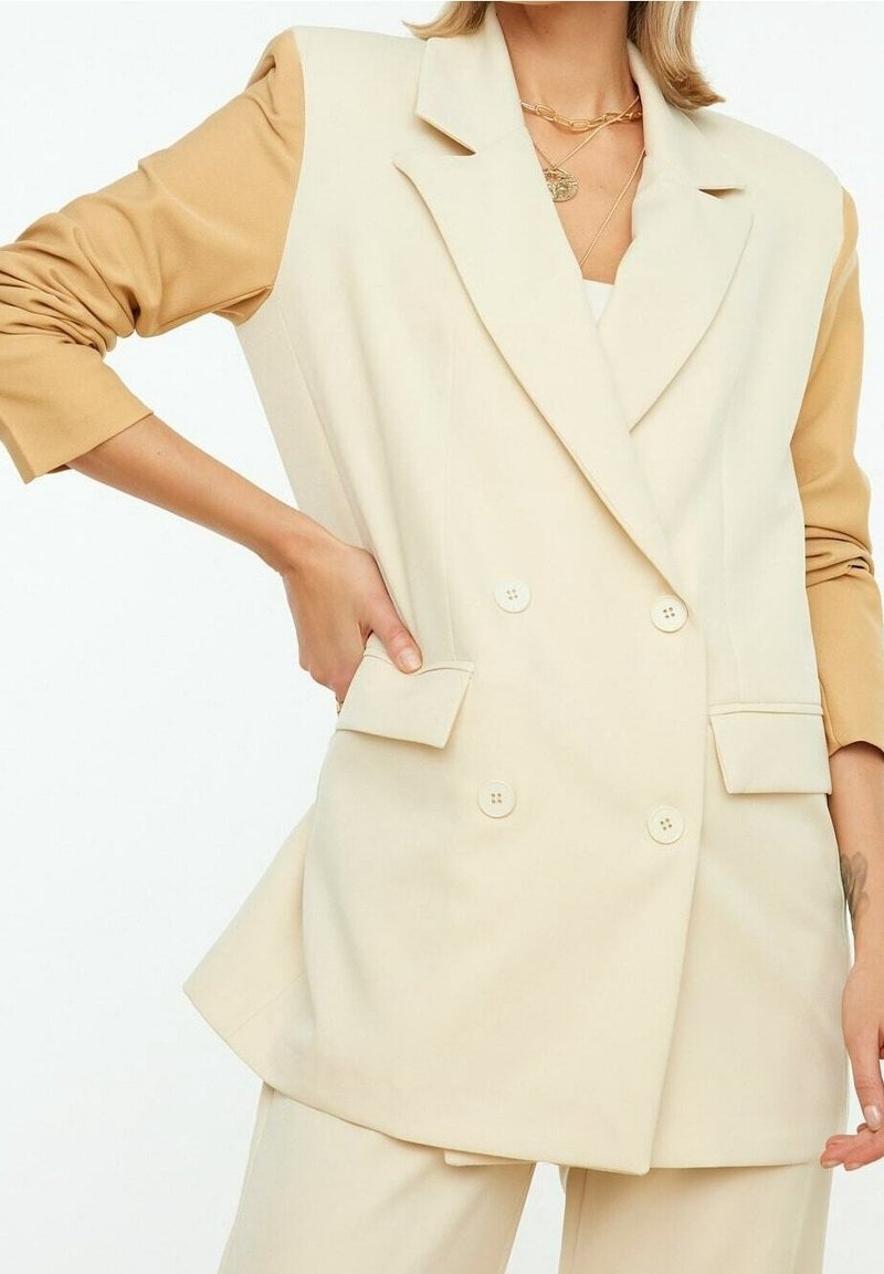 Beige doppelreihiger Blazer mit strukturierten Schultern, kontrastierenden tanfarbenen Ärmeln und taillierter Passform. Verfügt über Taschen vorne und Knopfdetails.