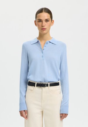 Femme aux cheveux attachés portant un polo bleu clair à manches longues rentré dans un pantalon beige taille haute avec une ceinture noire.