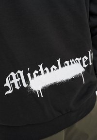Sudadera negra con la palabra "Michelangelo" en fuente gótica blanca, con un efecto de pintura en spray texturizado y un dobladillo acanalado.