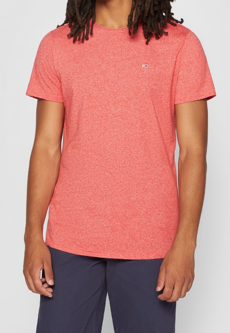 Homme portant un t-shirt corail chiné à manches courtes et col rond, ainsi qu'un pantalon bleu foncé, debout avec les bras détendus sur un fond uni.