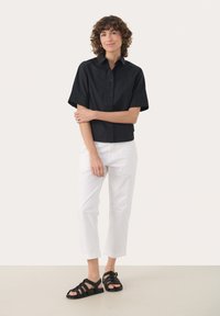 Černá košile s krátkým rukávem na knoflíky, bílé cropped kalhoty a černé sandály na platformě na modelce stojící na světlejším pozadí.