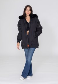 Zwarte bomberjack met een nep bont capuchon, ritssluiting, geribde mouwen en een oranje label. Gekenmerkt met een flared blauwe spijkerbroek en witte sneakers.