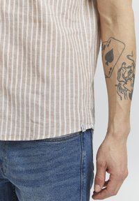 Chemise à manches courtes à rayures beiges en tissu léger ; présente de fines rayures blanches, un ourlet droit et une petite étiquette de marque sur le côté.